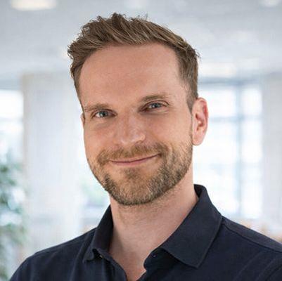 Porträt von Tobias Dreßler: Ein lächelnder Mann mit kurzen, hellbraunen Haaren und leichtem Bart schaut direkt in die Kamera. Er trägt ein dunkles Poloshirt und steht vor einem hellen, unscharfen Hintergrund, der wie ein modernes Büro wirkt